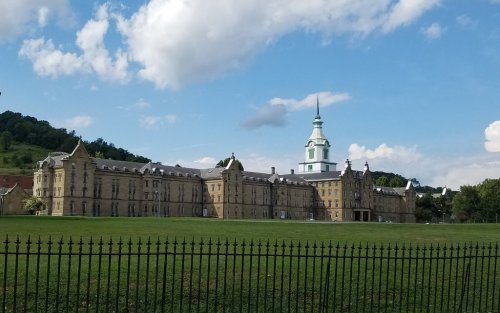 Historic building with a clock tower, surrounded by green grass and a black fence, under a blue sky with clouds.