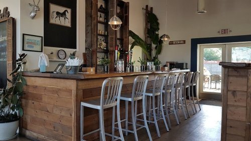 A rustic bar with metal stools, shelves of drinks, and large windows letting in natural light. Green plants add decor.