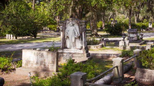 A serene cemetery scene featuring a stone monument with a statue and engraved gravestones surrounded by greenery.