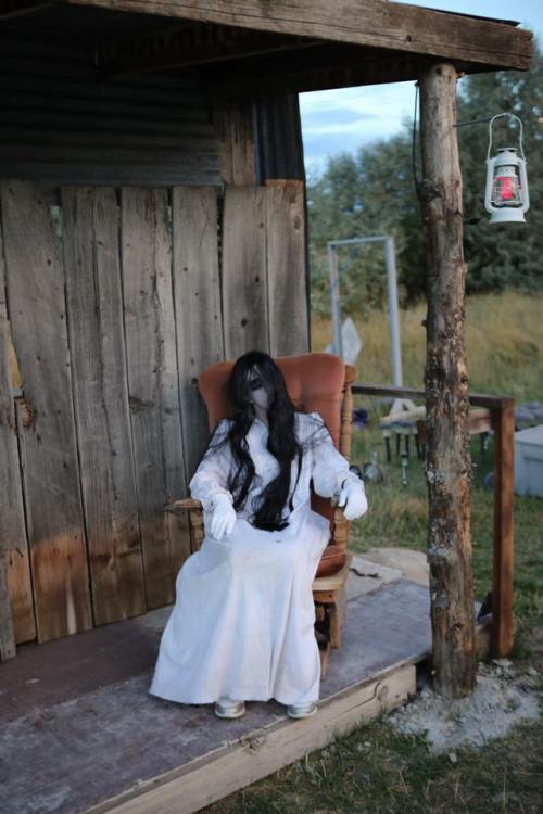 A creepy doll in a white dress with long black hair sits in a chair on a rustic porch, surrounded by a rural setting.