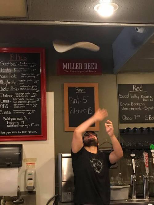 A person tosses pizza dough in the air at a pizzeria, with a menu and beer sign in the background.