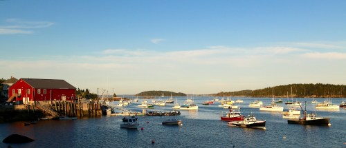 A serene harbor scene with a red building, numerous boats, and lush green islands under a clear blue sky.