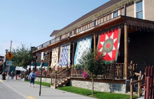 Colorful quilts hang from a wooden porch, with shops and trees lining the street under a clear blue sky.