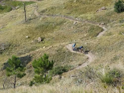 Two mountain bikers navigate a winding trail through grassy hills and rocky terrain.