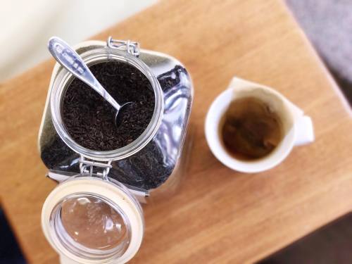 A glass jar filled with loose tea leaves next to a cup of brewed tea on a wooden table.