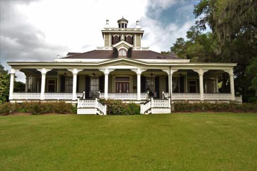 A large, historic white mansion with a wraparound porch, surrounded by green grass and trees under a cloudy sky.