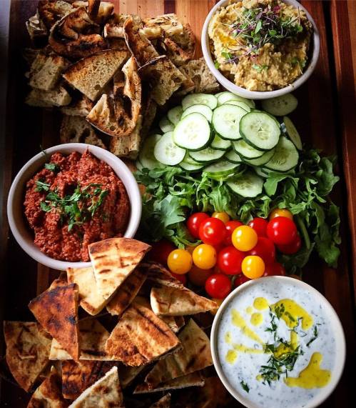 A wooden platter with pita bread, hummus, red dip, cucumber slices, cherry tomatoes, and a yogurt dip with herbs.