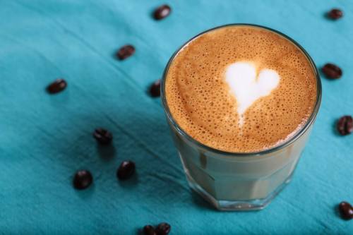 A glass of coffee with a heart-shaped foam design, surrounded by coffee beans on a turquoise background.