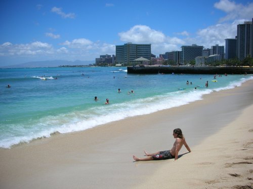 A person sits on a sandy beach, enjoying the ocean view with surfers and city buildings in the background.