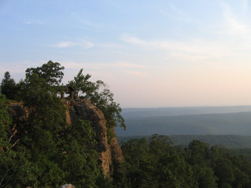 A scenic view of a rocky cliff with a small gazebo, surrounded by lush green trees and distant mountains under a clear sky.