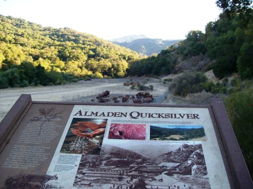 Sign about Almaden Quicksilver with historical images, overlooking a valley surrounded by green hills and mountains.