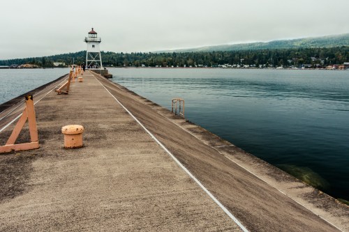 A lighthouse stands at the end of a long, concrete pier extending into calm waters, surrounded by trees and hills.