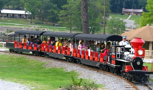 A colorful miniature train with passengers rides along a track in a scenic outdoor setting.