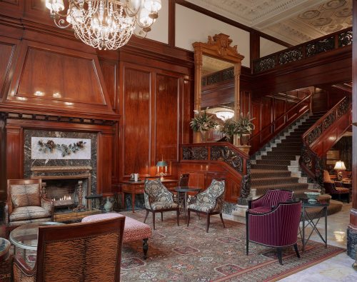 Elegant hotel lobby featuring wooden paneling, a chandelier, plush seating, and a grand staircase.