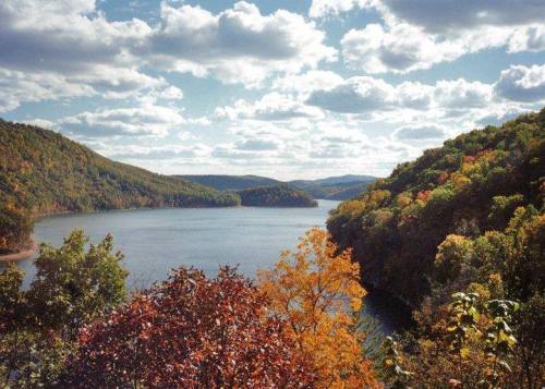 A scenic view of a lake surrounded by colorful autumn foliage and rolling hills under a partly cloudy sky.