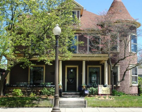 A charming two-story house with a turret, surrounded by trees and a lamp post, featuring a welcoming front porch.