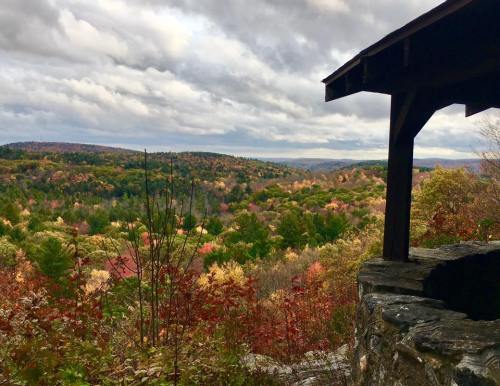 Scenic view of colorful autumn foliage across rolling hills under a cloudy sky, framed by a rustic wooden structure.