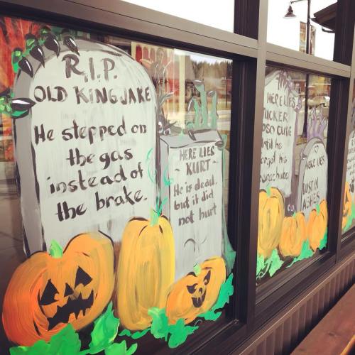 Window display featuring Halloween-themed artwork with tombstones, pumpkins, and humorous epitaphs.