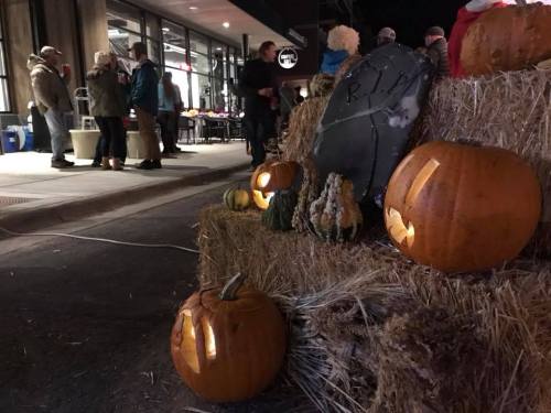 A festive scene with carved pumpkins and gourds on hay bales, with people mingling in the background at night.