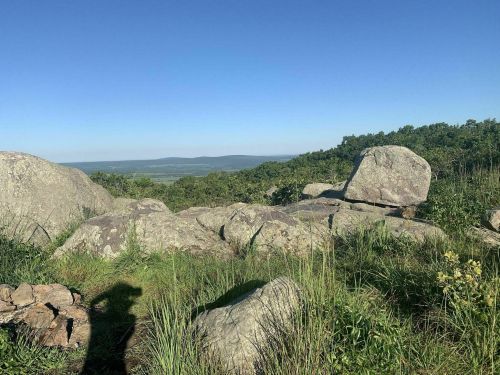 A scenic view of large rocks and green grass, overlooking a valley under a clear blue sky.