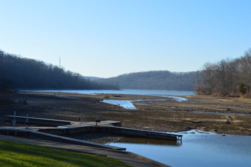 A serene riverbank scene with a dry shoreline, wooden docks, and distant hills under a clear blue sky.