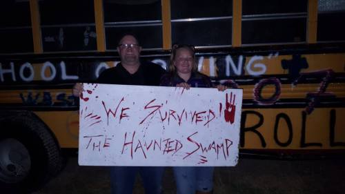 A man and woman hold a sign reading "We Survived the Haunted Swamp" in front of a yellow school bus at night.