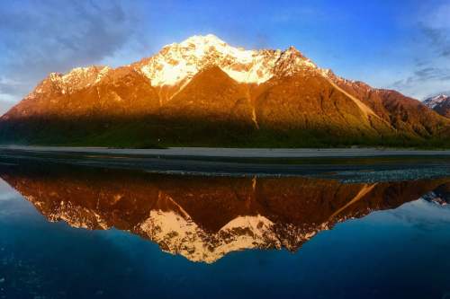 A majestic mountain with snow-capped peaks reflects in a calm lake at sunset, surrounded by lush greenery.