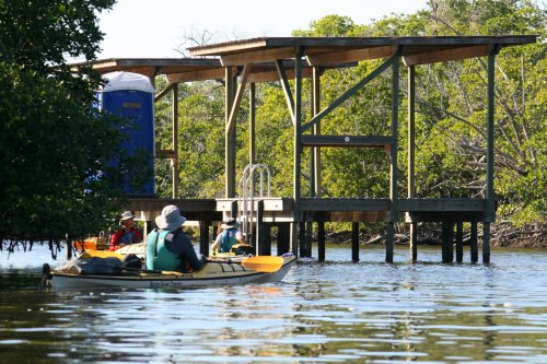 Two kayakers approach a wooden dock surrounded by lush greenery, with a portable restroom visible on the structure.