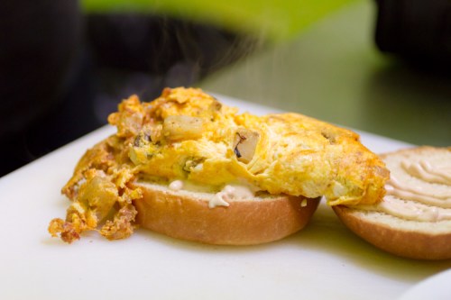 A warm omelet with vegetables sits on a bun, topped with a creamy spread, on a cutting board. Steam rises from the dish.