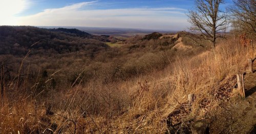 A panoramic view of a valley surrounded by rolling hills and sparse trees under a clear blue sky.