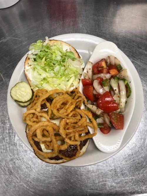 A plate with a burger bun topped with lettuce, crispy onion rings, a side of fresh tomato and onion salad, and a pickle.