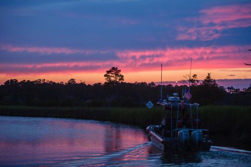 A boat glides through a calm waterway at sunset, with vibrant pink and purple skies reflecting on the water.