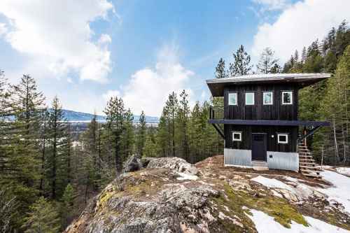 A modern black cabin on a rocky outcrop, surrounded by pine trees and mountains under a partly cloudy sky.