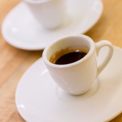 A small white espresso cup filled with dark coffee on a white saucer, with another cup blurred in the background.