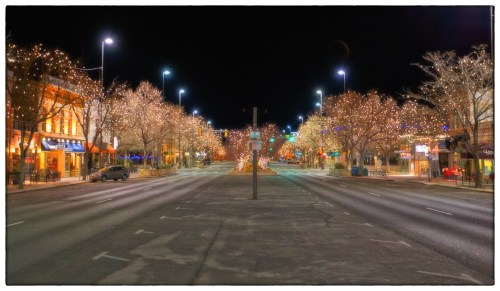 A quiet, empty street at night, lined with trees adorned with twinkling lights and illuminated buildings.