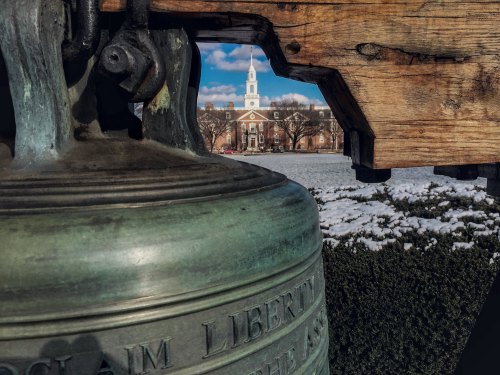 A close-up of a bell with a wooden frame, framed by a snowy landscape and a church steeple in the background.