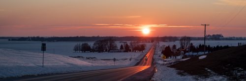 A scenic winter road at sunset, with snow-covered fields and distant trees under a colorful sky.