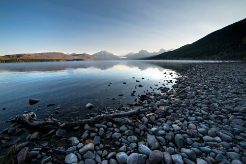 A serene lake scene with smooth stones along the shore, mountains in the background, and a calm, reflective water surface.