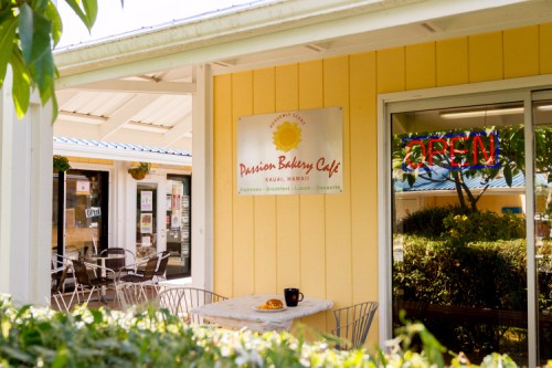 Exterior of Passion Bakery Cafe in Kauai, Hawaii, featuring a sign and an "OPEN" neon light. Tables are visible outside.