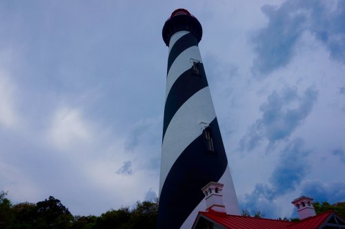 A tall, striped lighthouse against a cloudy sky, with a red roof and surrounding greenery.