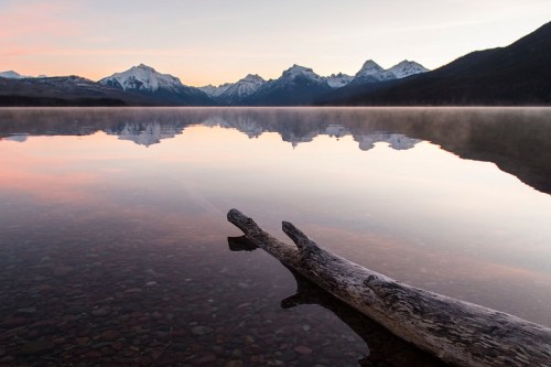 A calm lake at sunrise, reflecting snow-capped mountains and a log on the shore, creating a serene landscape.