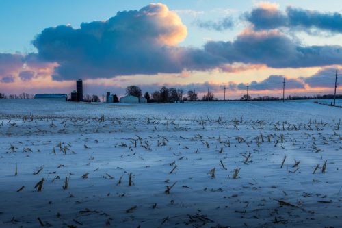 A snowy field at sunset, with clouds and a distant farm silhouette under a colorful sky.