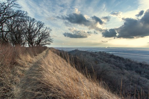 A scenic view of a grassy ridge overlooking a valley with trees and a cloudy sky in the background.