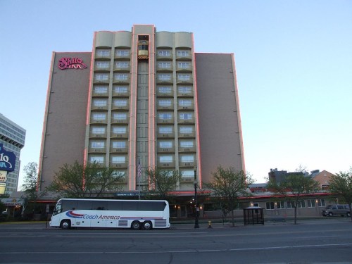 A tall hotel building with a bus parked in front, surrounded by trees and a clear blue sky.