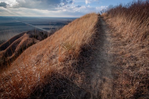 A winding dirt path through dry grass on a hillside, overlooking a vast, open landscape under a cloudy sky.