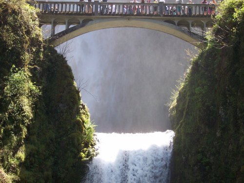 A stone bridge spans a waterfall, surrounded by lush greenery and mist rising from the cascading water below.