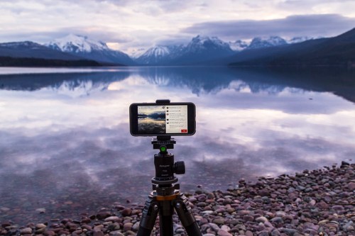 A smartphone on a tripod captures a scenic lake and mountains, reflecting the cloudy sky in the calm water.