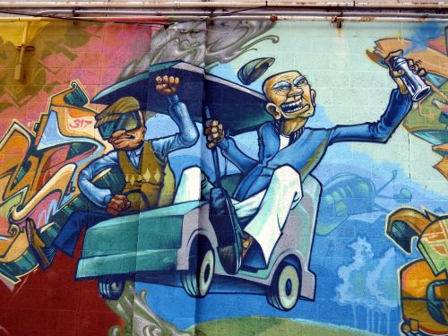 Colorful mural depicting two cartoonish characters in a car, one wearing sunglasses and the other holding a spray can.
