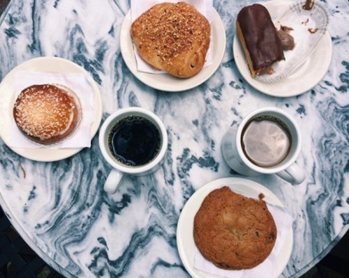 A marble table with two cups of coffee, a cookie, a pastry, and a chocolate-covered treat on white plates.