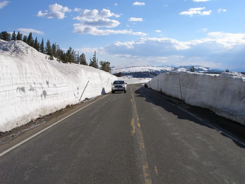 A car drives on a snow-lined road with tall snowbanks on either side under a blue sky with scattered clouds.